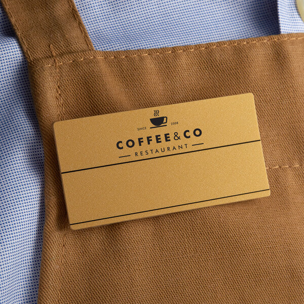 A brown apron with a Cawley customizable gold plastic rectangle nametag on a counter in a coffee shop.
