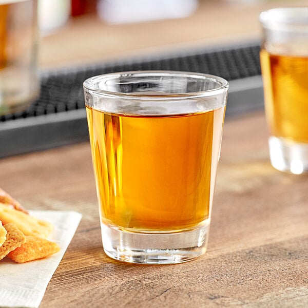 An Acopa shot glass filled with amber liquid on a table with a plate of food.