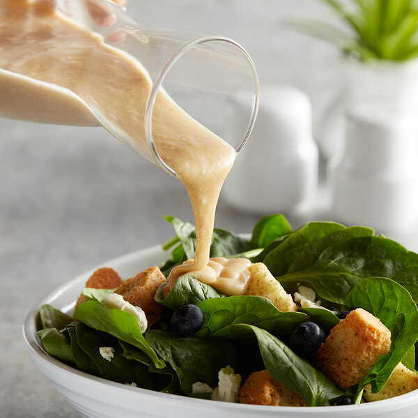 A bowl of spinach salad with blueberries, croutons, and AAK Select Honey Mustard Dressing being poured over it.