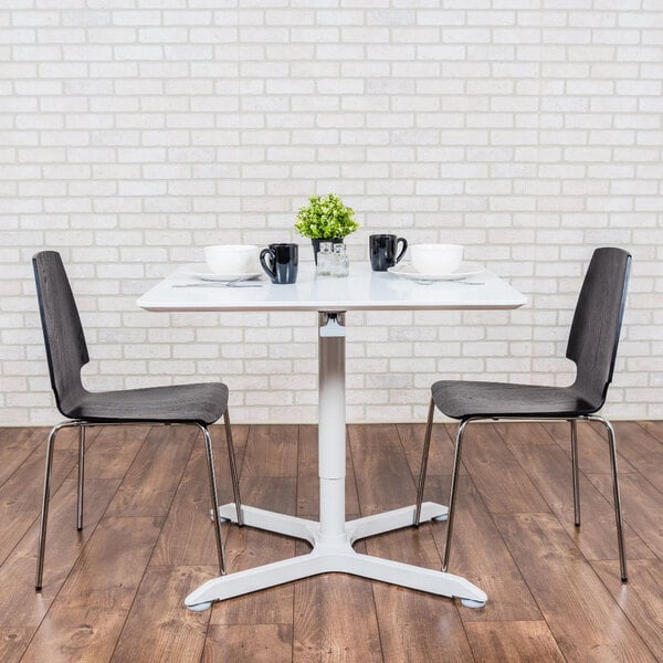 A white Luxor square pneumatic adjustable height cafe table with two black chairs on a restaurant dining area.