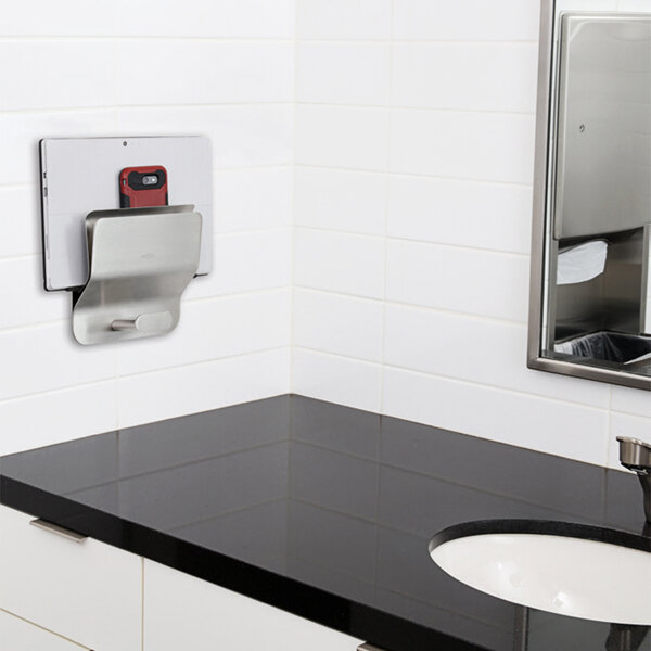 A Bobrick stainless steel mobile device holder on a wall above a sink.