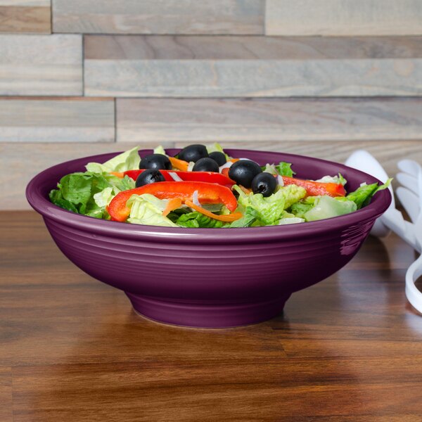 A purple Fiesta pedestal serving bowl with salad and green vegetables in it.