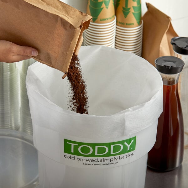 A bag of coffee being poured into a Toddy coffee container with a paper filter.