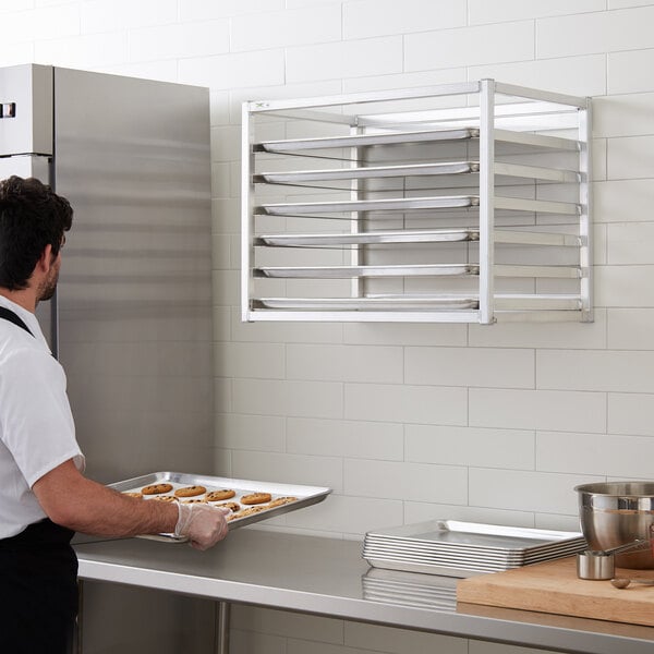 A man wearing a white shirt and gloves using a Regency wall mounted sheet pan rack to hold a tray of cookies.
