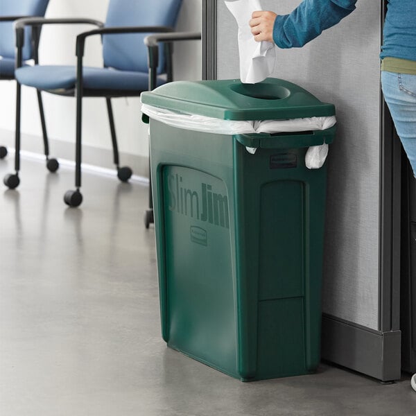 A woman putting paper in a green Rubbermaid Slim Jim trash can.
