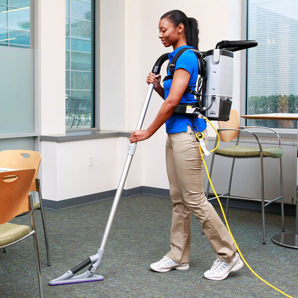 A woman using a ProTeam backpack vacuum to clean a chair in a corporate office cafeteria.