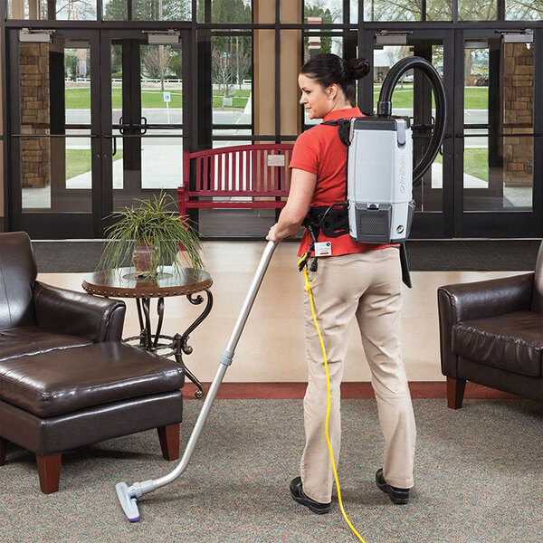 A woman in a red shirt using a ProTeam backpack vacuum to clean a room with brown leather chairs.