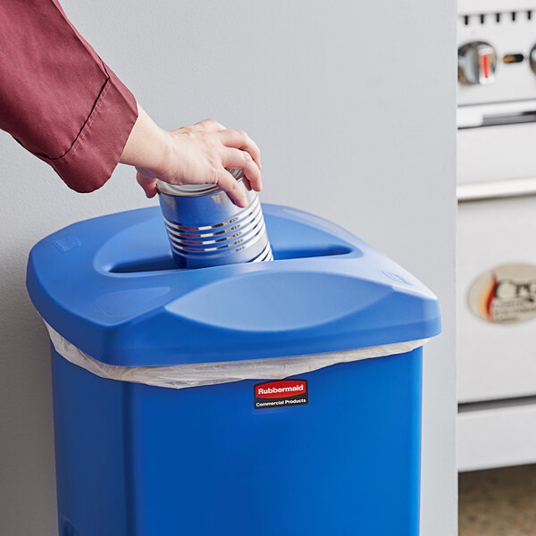 A hand putting a blue can with a mixed recycling lid into a blue Rubbermaid commercial trash can.