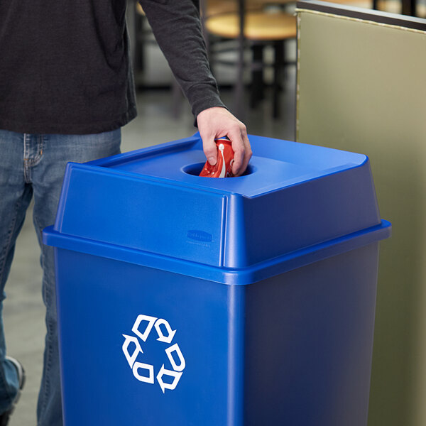 A person putting a can into a blue Rubbermaid Untouchable recycling bin.
