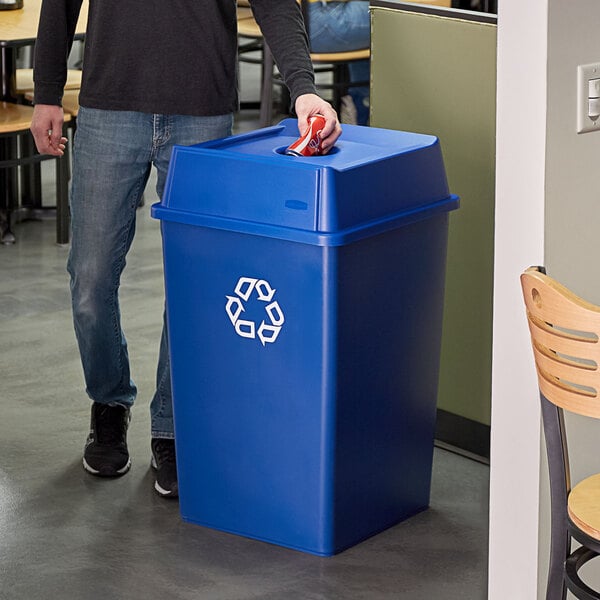 A person standing next to a Rubbermaid blue recycle bin with a bottle/can hole lid.