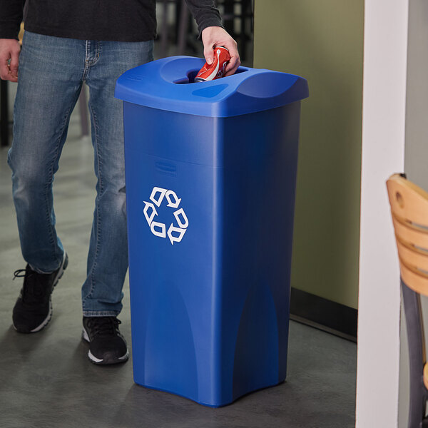 A man standing next to a Rubbermaid blue recycle bin with a mixed recycle slot lid.
