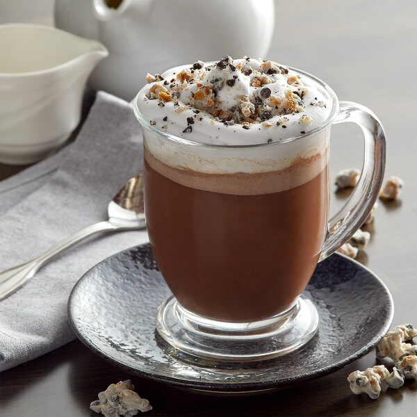 A glass cup of HERSHEY'S Cookies 'n' Creme hot chocolate with whipped cream and crumbs.