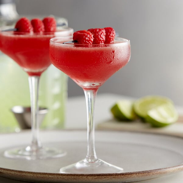 Two Arcoroc tall coupe cocktail glasses filled with raspberry margaritas and garnished with raspberries on a table.