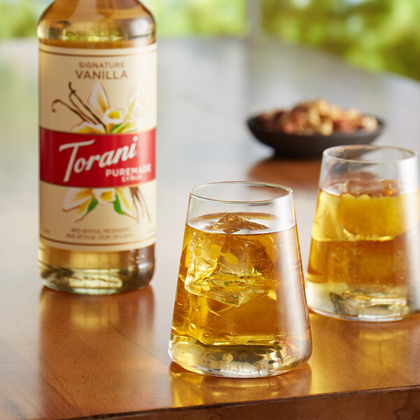 A close up of a Torani Puremade Signature Vanilla flavoring syrup bottle next to two glasses of vanilla-flavored liquid.