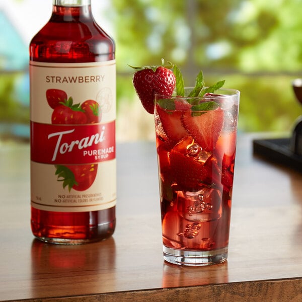 A glass of red liquid with ice and strawberries next to a bottle of Torani Puremade Strawberry flavoring syrup.