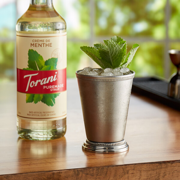 A silver cup with ice and mint leaves next to a bottle of Torani Puremade Creme de Menthe syrup.