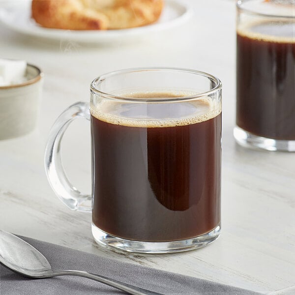 Two Acopa clear glass coffee mugs of coffee with a spoon on a napkin.