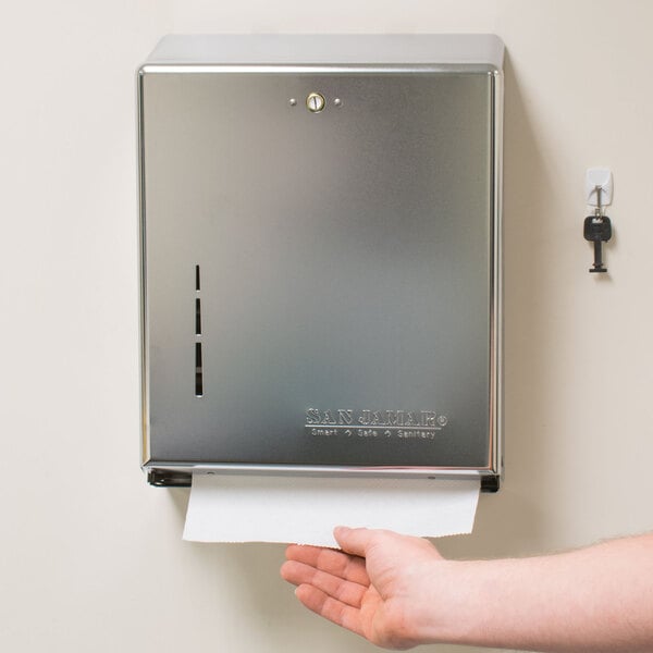 A hand using a San Jamar Chrome C-Fold / Multi-Fold Towel Dispenser on a wall.