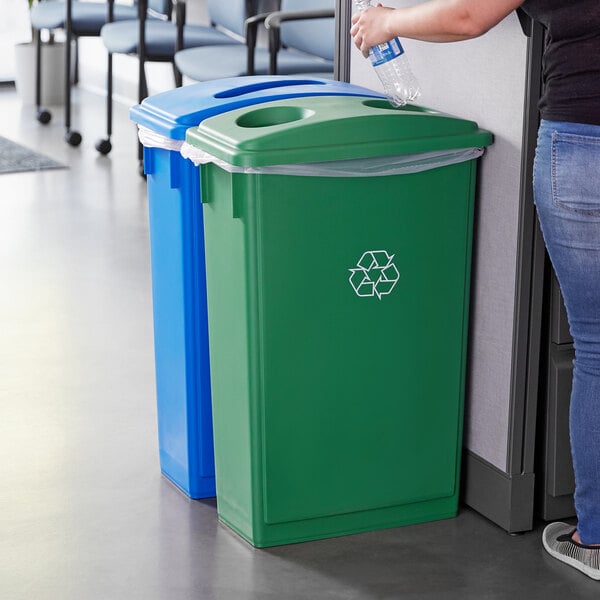 A woman standing next to a Lavex green recycle bin with a green bottle in her hand.