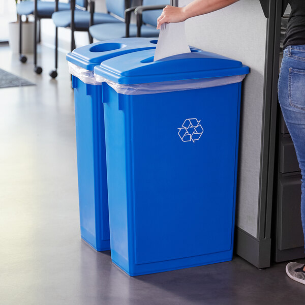 A woman standing next to a Lavex blue recycle bin with paper, bottle, and can lids.
