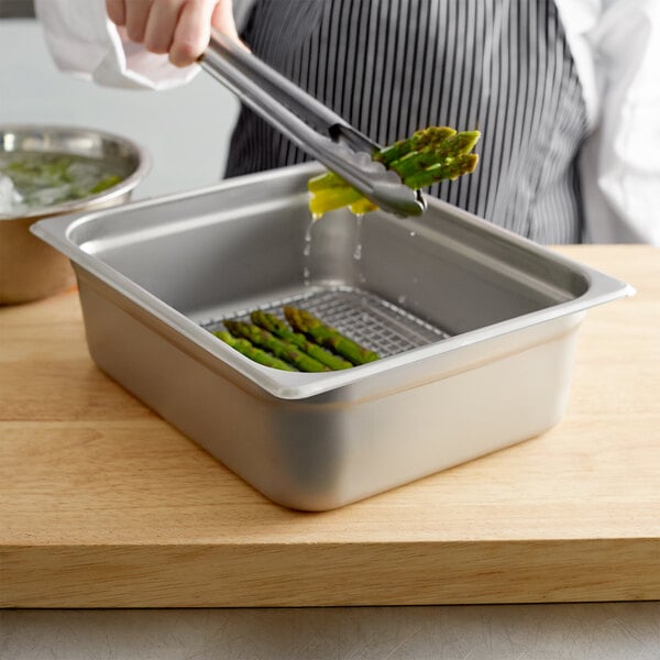 A person using tongs to remove asparagus from a Choice 1/2 size stainless steel pan.