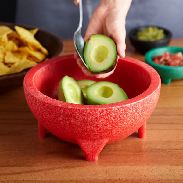 A hand holding half of an avocado being cut up in a red Molcajete bowl.