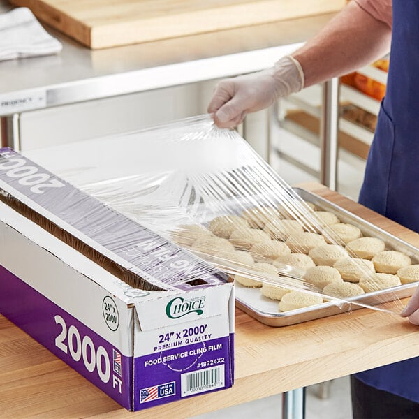 A person in a blue shirt using Choice Foodservice Film with a serrated cutter to cover a tray of cookies.