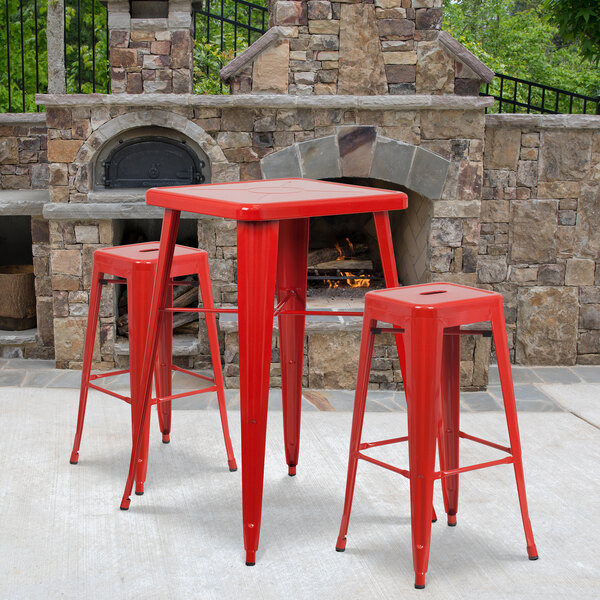 A red metal table and bar stools on an outdoor patio in front of a brick fireplace.