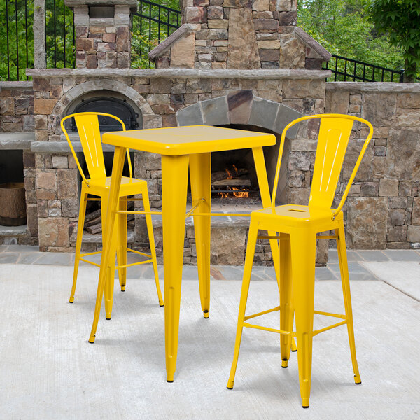 A Flash Furniture yellow metal bar height table with two yellow chairs on an outdoor patio next to a stone fireplace.