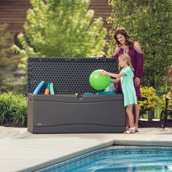 A woman and a girl sit by a pool on an outdoor patio with a Lifetime outdoor storage box.