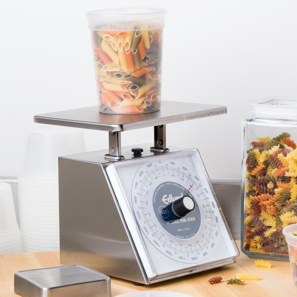 An Edlund Four Star portion scale on a counter with a plastic container full of pasta being weighed.