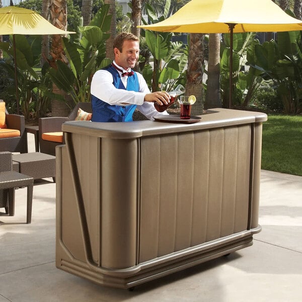 A man in a blue vest standing behind a Cambro portable bar with a drink.