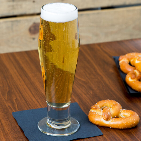 A Libbey footed pilsner glass of beer on a table with a pretzel.
