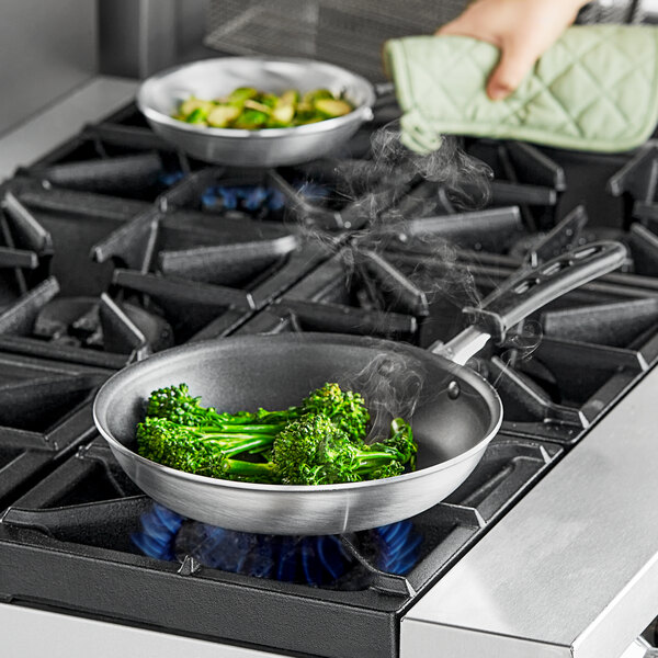 A hand using a green Vollrath Wear-Ever frying pan to cook broccoli on a stove.