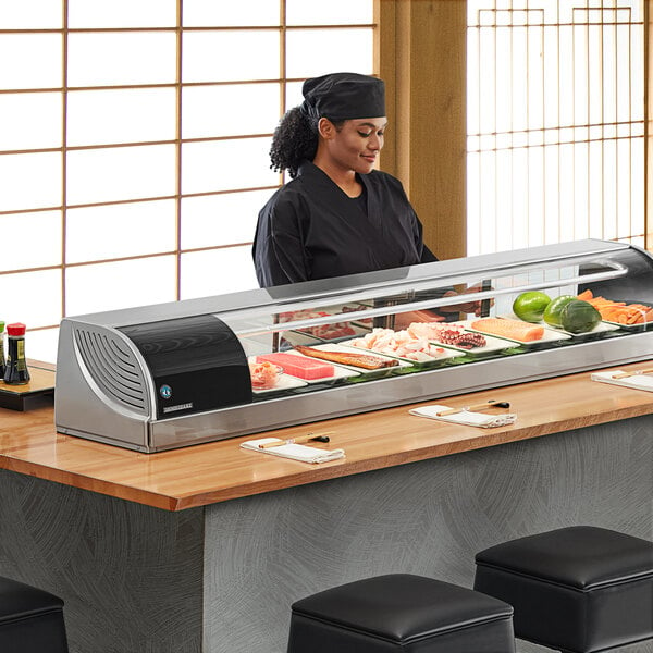 A woman standing behind a Hoshizaki refrigerated sushi display case.