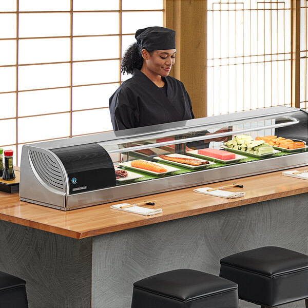 A woman standing behind a refrigerated sushi display case full of sushi.