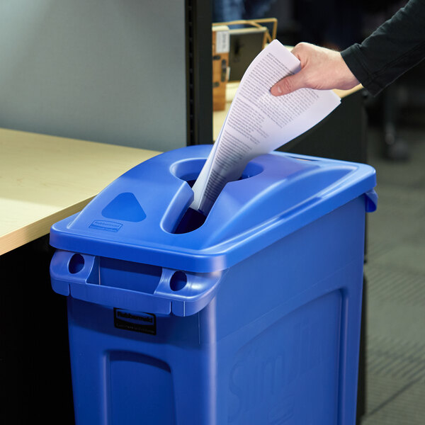 A hand putting a piece of paper into a blue Rubbermaid recycling bin with a mixed recycle slot.