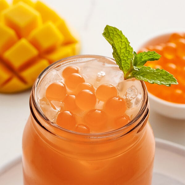 A glass of orange liquid with Bossen Mango Bursting Boba and mint leaves on a table.