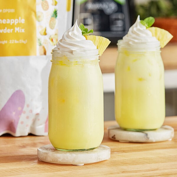 Two glass jars filled with yellow pineapple drinks with whipped cream on top.