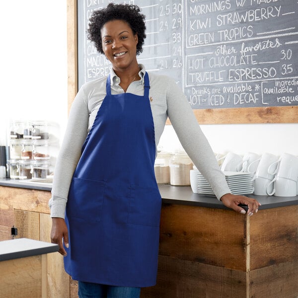 A smiling woman wearing a Choice royal blue customizable bib apron standing behind a counter.