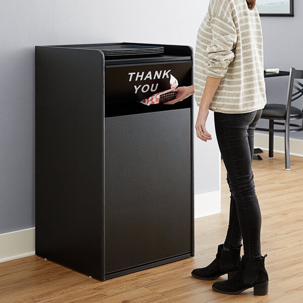 A woman standing next to a black rectangular Lancaster Table & Seating waste receptacle enclosure with a "THANK YOU" sign on the door.