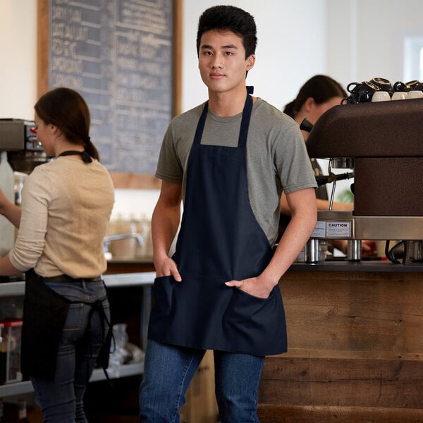 A man in a navy blue Choice bib apron with three pockets standing in a professional kitchen.