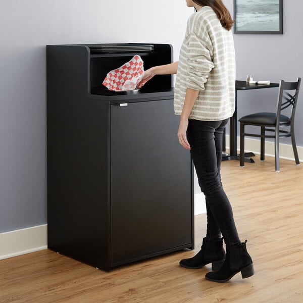 A woman standing in front of a black Lancaster Table & Seating waste receptacle enclosure.