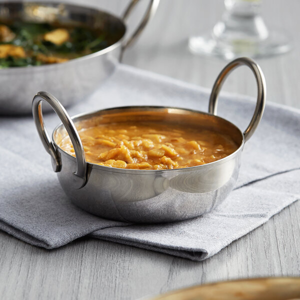 A Vollrath stainless steel balti dish filled with soup on a table.