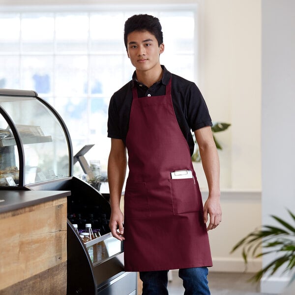 A man wearing a burgundy standard bib apron with 2 pockets standing at a counter.