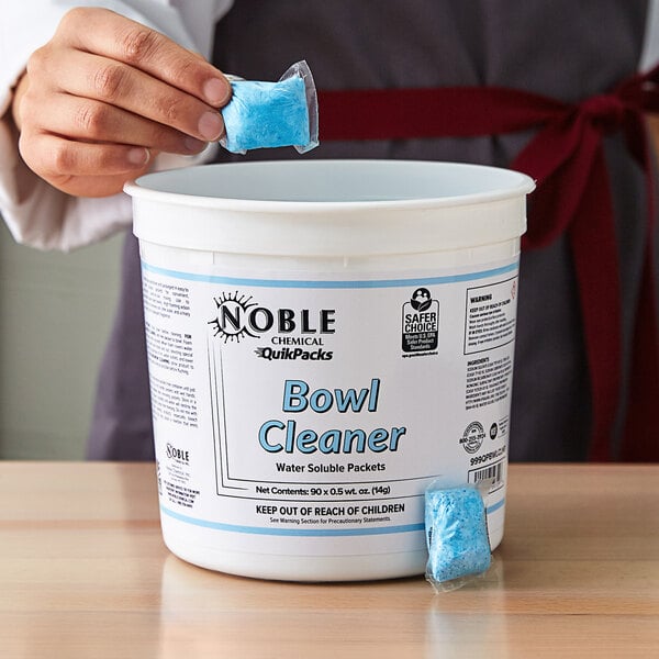 A person putting a blue Noble Chemical QuikPacks bowl cleaner in a white bucket.