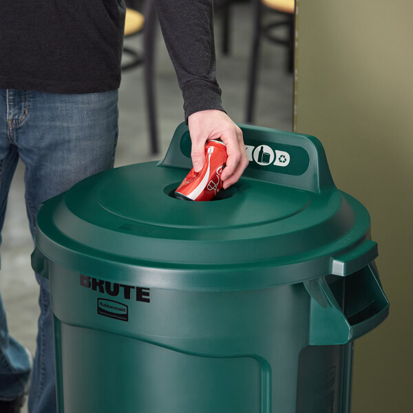 A person putting a can into a green Rubbermaid recycling bin with a lid.