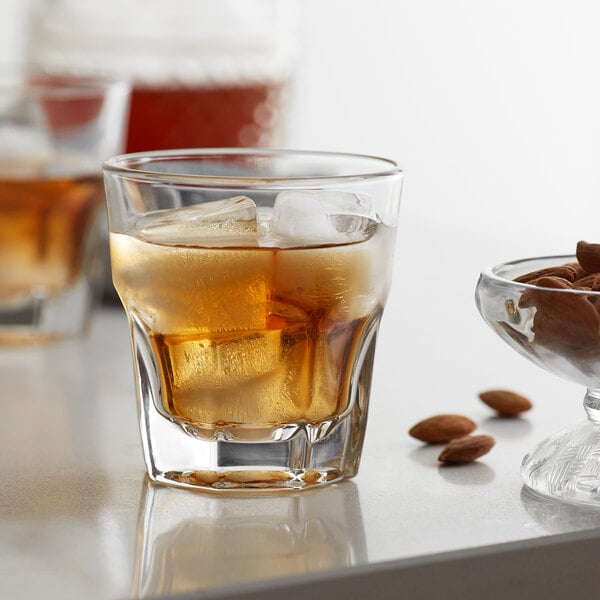 A close-up of a Libbey Gibraltar Rocks Glass with ice and brown liquid, next to a bowl of almonds.