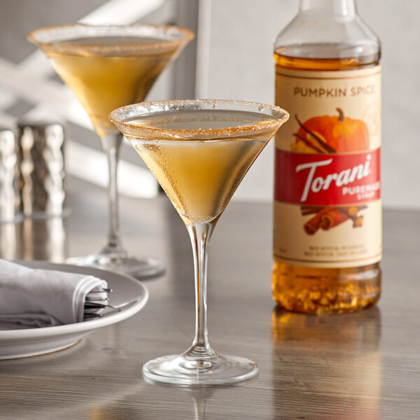 A martini glass filled with brown liquid next to a bottle of Torani Pumpkin Spice syrup.