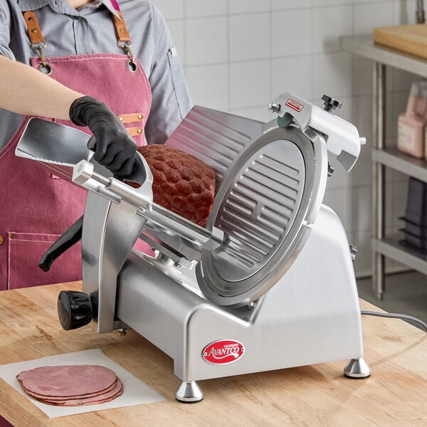 A person wearing a pink apron and black gloves is using a large, silver meat slicer to slice a piece of ham, with several slices already placed on a piece of paper on the wooden counter.
